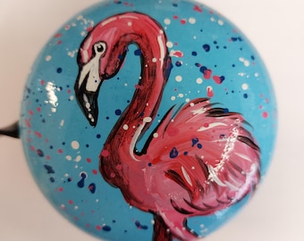 Peut inclure: Une sonnette de vélo ronde peinte à la main, ornée d'un flamant rose sur fond bleu clair. Le flamant a un bec blanc et est entouré d'éclaboussures de peinture colorées. Un accessoire décoratif pour vélos.