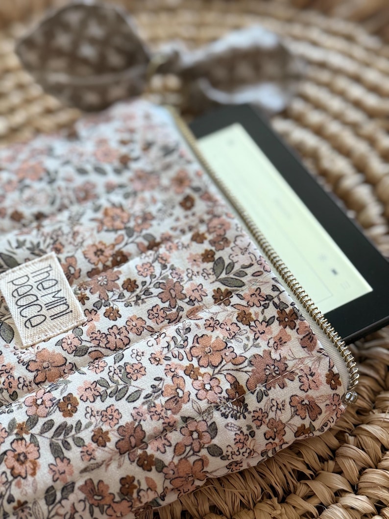 May include: A floral print e-reader case with a gold zipper, holding an e-reader. The case features a cream background with a pattern of small pink and brown flowers and green leaves. The label reads "the mini peace".