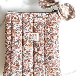 May include: A floral print quilted pouch with a matching bow-shaped keychain. The pouch features a white background with a pattern of pink and brown flowers and green leaves. The bow is a light brown color with a cross pattern. The text "The Mini Peace" is on the pouch.