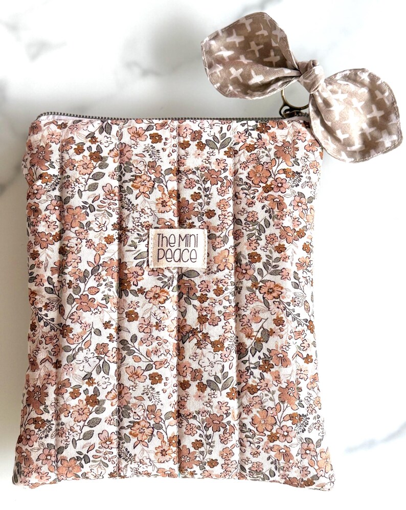 May include: A floral print quilted pouch with a matching bow-shaped keychain. The pouch features a white background with a pattern of pink and brown flowers and green leaves. The bow is a light brown color with a cross pattern. The text "The Mini Peace" is on the pouch.