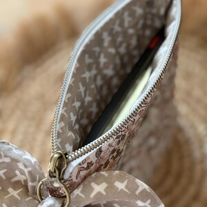 May include: A floral print pouch with a gold zipper and a matching bow. The interior lining features a pattern of white crosses on a beige background. A black object is partially visible inside the pouch.