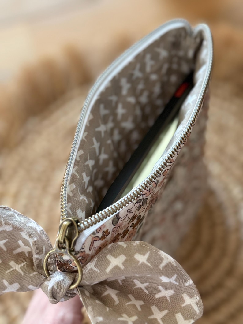 May include: A floral print pouch with a gold zipper and a matching bow. The interior lining features a pattern of white crosses on a beige background. A black object is partially visible inside the pouch.