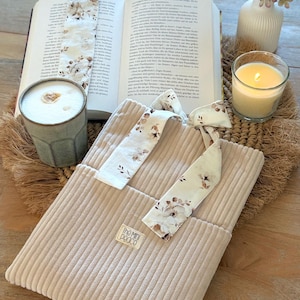 Könnte beinhalten: Eine beigefarbene Buchhülle aus Cord mit einem weißen Stoffband mit braunem Blumenmuster, das zu einer Schleife gebunden ist. Ein kleines rechteckiges Etikett mit der Aufschrift "the mini peace" ist auf die Vorderseite der gerippten Hülle genäht.