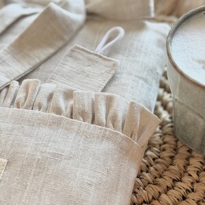 May include: A close-up of a light beige linen robe with a ruffled edge and a matching belt. The robe is folded on a woven mat, with a mug of foamy liquid in the background. The robe has a textured appearance.