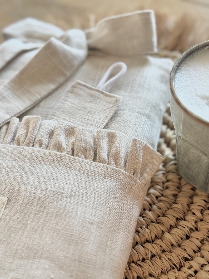 May include: A close-up of a light beige linen robe with a ruffled edge and a matching belt. The robe is folded on a woven mat, with a mug of foamy liquid in the background. The robe has a textured appearance.