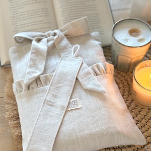 May include: A beige linen book sleeve with a matching bookmark, tied with a bow. The bookmark has a small tag that reads "mini peace". The sleeve is next to an open book, a candle, and a mug.