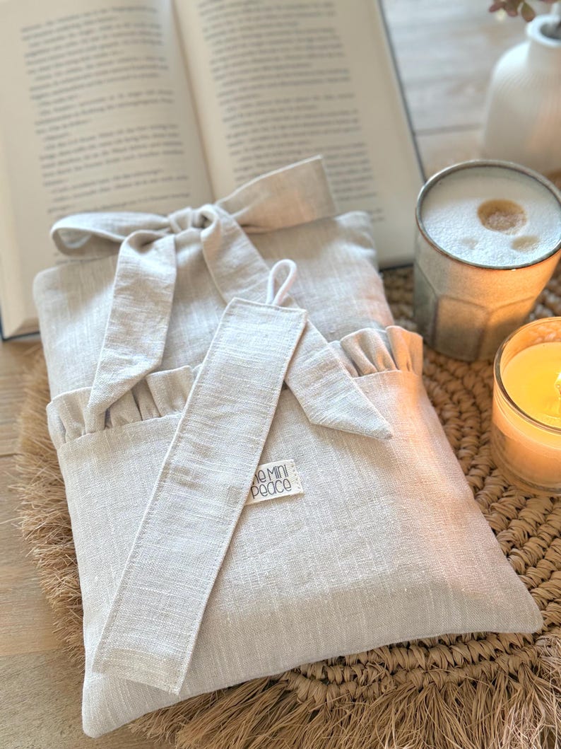 May include: A beige linen book sleeve with a matching bookmark, tied with a bow. The bookmark has a small tag that reads "mini peace". The sleeve is next to an open book, a candle, and a mug.