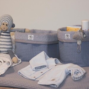 Puede incluir: Dos cestas de almacenamiento de tejido de gofre gris con un juguete de mono de crochet a rayas gris y blanco, un body de beb&eacute; blanco y una manta blanca. Las cestas tienen un forro de tela gris y una etiqueta blanca con la letra "P".