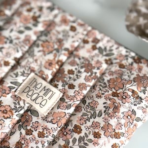 May include: Close-up of a floral-patterned fabric with a quilted design. The fabric features a cream background with a repeating pattern of small, peach and brown flowers and green leaves. A small rectangular tag with the text "the mini peace" is sewn onto the fabric.
