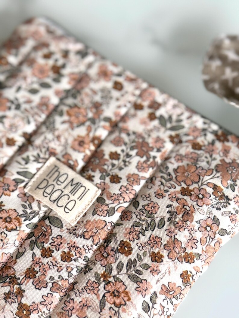 May include: Close-up of a floral-patterned fabric with a quilted design. The fabric features a cream background with a repeating pattern of small, peach and brown flowers and green leaves. A small rectangular tag with the text "the mini peace" is sewn onto the fabric.