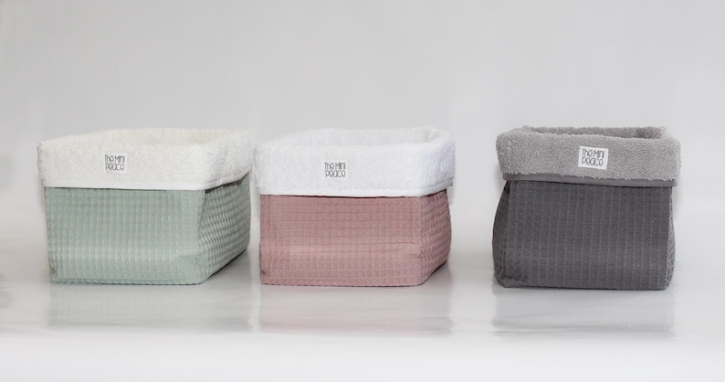 Puede incluir: Tres cestas de almacenamiento hechas de tela suave y texturizada. Las cestas son de color verde, rosa y gris. Cada cesta tiene una parte superior de tela blanca con el texto "TOMMI TOCCO" impreso en ella.