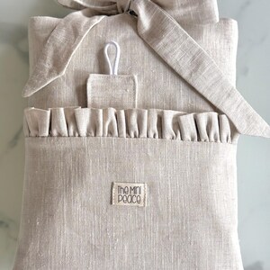 May include: A beige linen pouch with a ruffled pocket and a bow closure. The pouch has a small rectangular tag that reads "The Mini Peace". A small loop is attached to the top of the pouch.