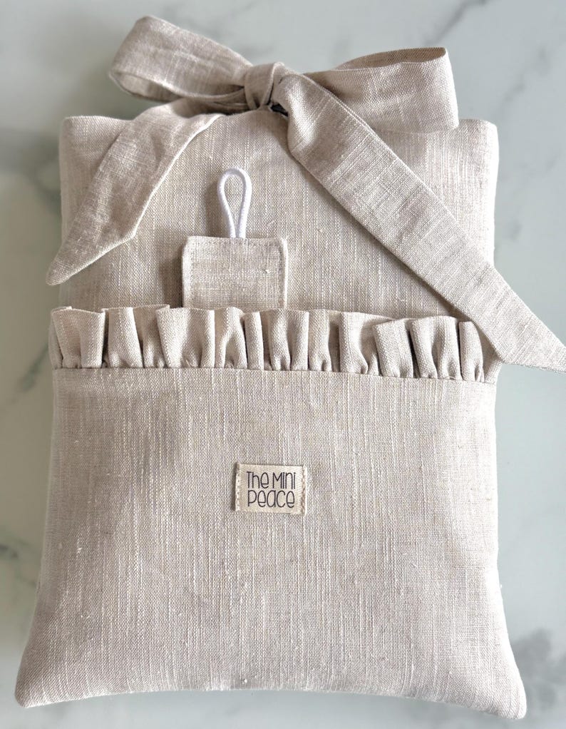 May include: A beige linen pouch with a ruffled pocket and a bow closure. The pouch has a small rectangular tag that reads "The Mini Peace". A small loop is attached to the top of the pouch.