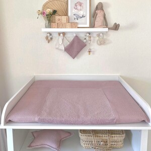 Può includere: Un fasciatoio bianco con un fasciatoio rosa a trama waffle. Una mensola sopra contiene un quadro incorniciato, blocchi di legno con la scritta "OH BABY" e oggetti decorativi. Un cuscino a forma di stella e un cestino in vimini sono sulla mensola sottostante.