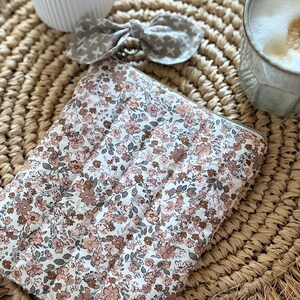 May include: A floral-patterned quilted pouch with a zipper closure, featuring a white background and small pink and brown flowers. A matching bow with a cross pattern and a ceramic vase are also visible.