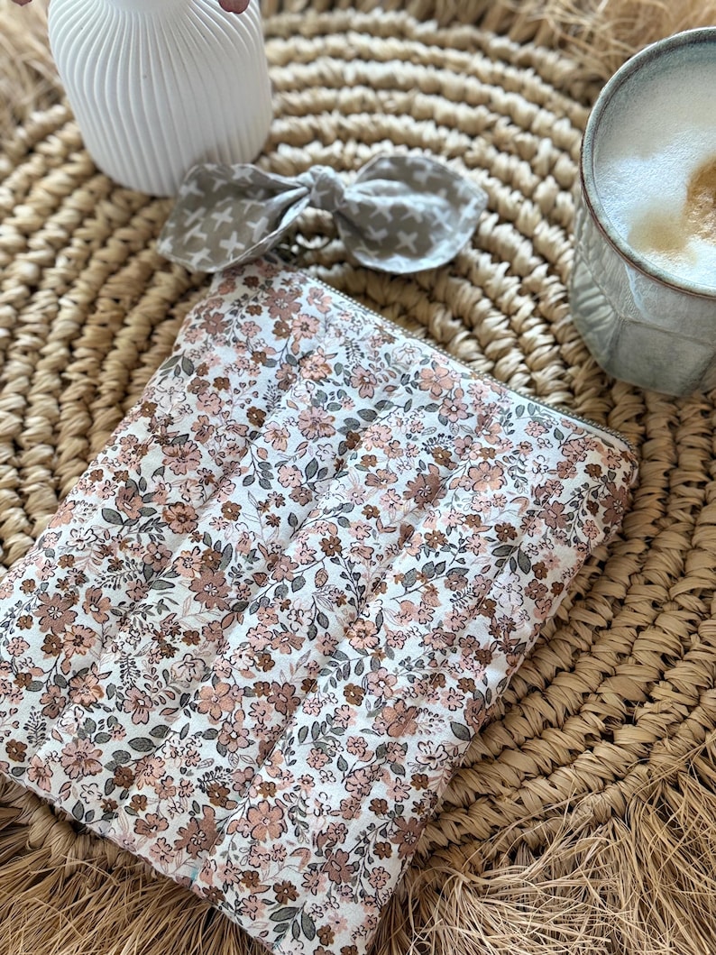 May include: A floral-patterned quilted pouch with a zipper closure, featuring a white background and small pink and brown flowers. A matching bow with a cross pattern and a ceramic vase are also visible.