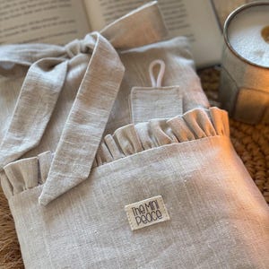 May include: A light beige linen book sleeve with a ruffled pocket and a matching tie closure. The label reads "The Mini Peace". The sleeve is designed to protect books and has a small loop for hanging.
