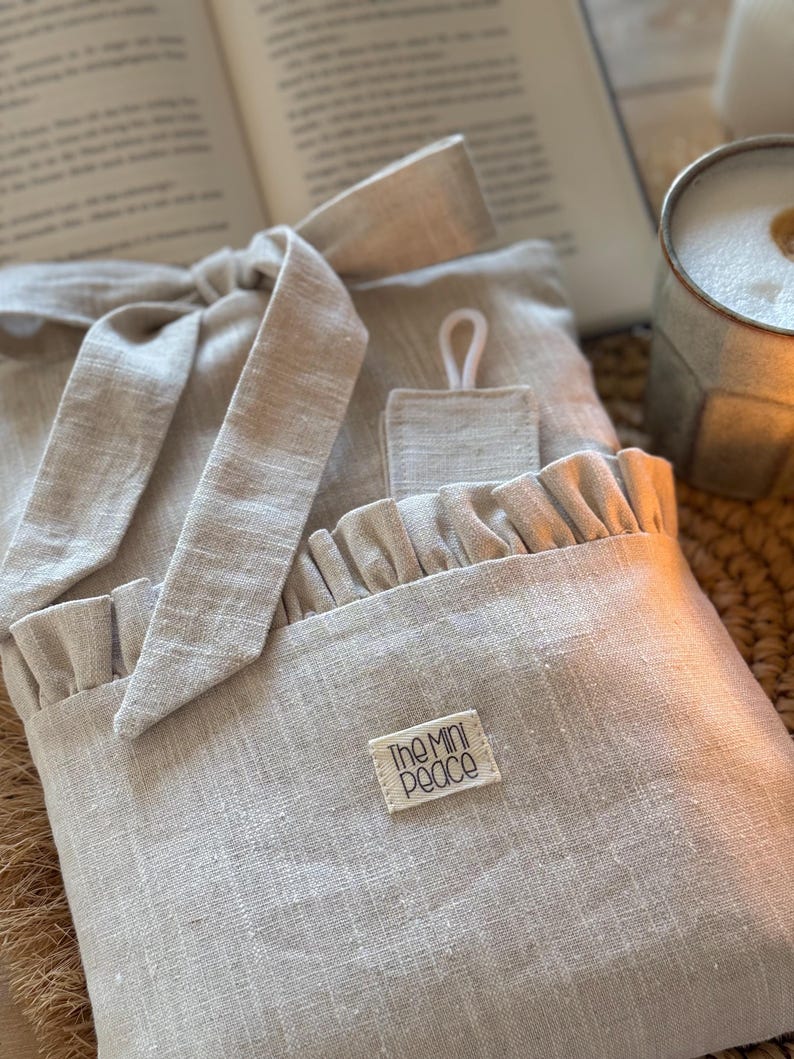 May include: A light beige linen book sleeve with a ruffled pocket and a matching tie closure. The label reads "The Mini Peace". The sleeve is designed to protect books and has a small loop for hanging.