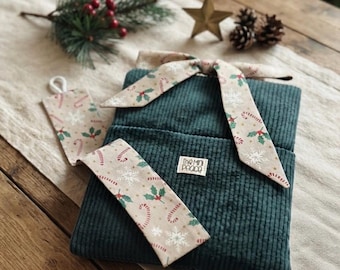May include: A dark green corduroy hot water bottle cover featuring a beige fabric tie with a Christmas pattern of candy canes, holly leaves, and snowflakes. A matching rectangular tag with the text The Mini Peace is attached to the cover.