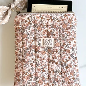 E-Reader Hülle  mit floralem Muster und Reißverschluss, die rosa und braune Blumen auf cremefarbenem Hintergrund zeigt. Die Hülle hat ein kleines Etikett mit der Aufschrift "The Mini Peace". Eine passende Schleife ist am Reißverschluss befestigt.