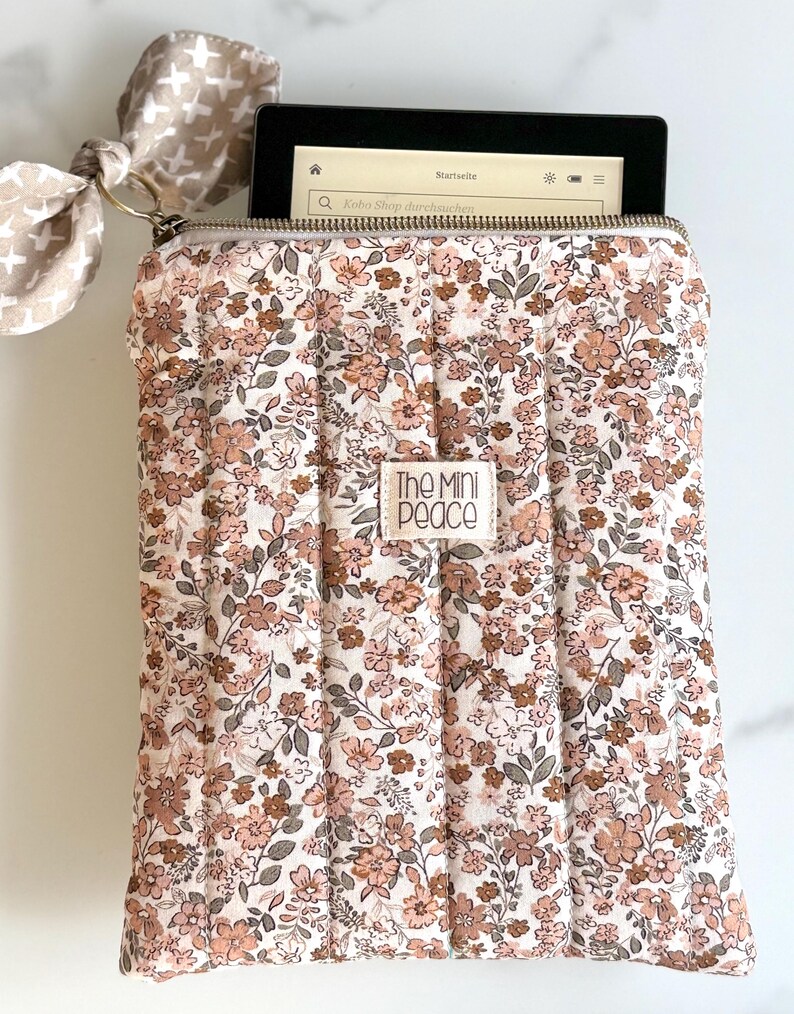 E-Reader Hülle  mit floralem Muster und Reißverschluss, die rosa und braune Blumen auf cremefarbenem Hintergrund zeigt. Die Hülle hat ein kleines Etikett mit der Aufschrift "The Mini Peace". Eine passende Schleife ist am Reißverschluss befestigt.