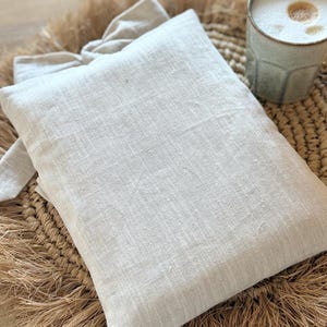 May include: A light beige linen pillow with a matching tie, resting on a woven straw mat. A light green ceramic mug filled with a frothy beverage sits nearby. The scene evokes a sense of comfort and relaxation.