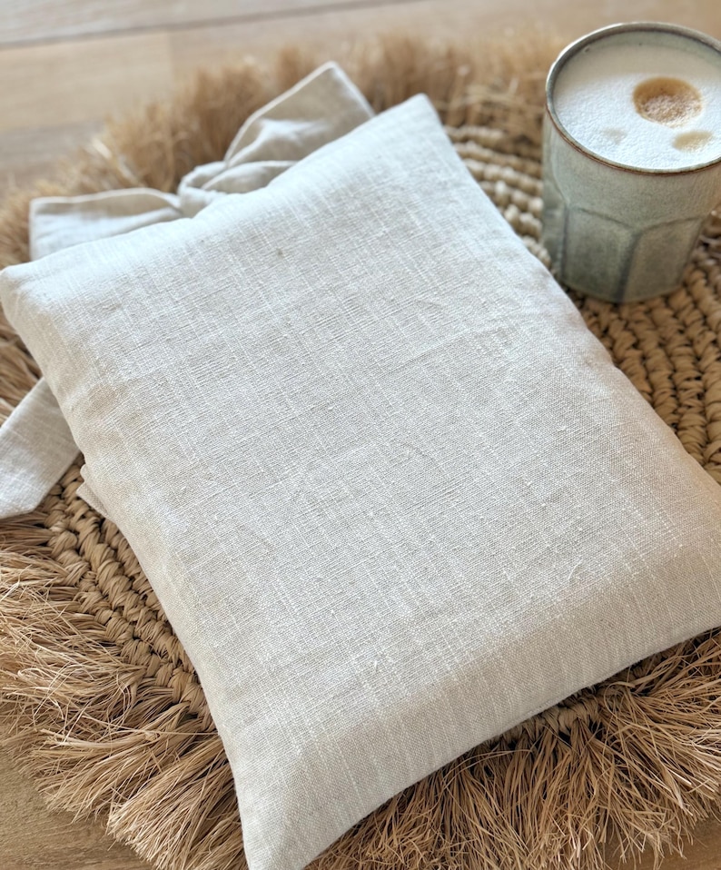 May include: A light beige linen pillow with a matching tie, resting on a woven straw mat. A light green ceramic mug filled with a frothy beverage sits nearby. The scene evokes a sense of comfort and relaxation.