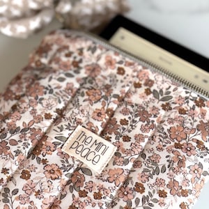 May include: A floral-patterned tablet sleeve with a zipper closure. The sleeve features a quilted design with a cream background and a pattern of pink and brown flowers and green leaves. The label reads "The Mini Peace".