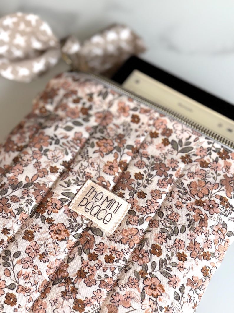 May include: A floral-patterned tablet sleeve with a zipper closure. The sleeve features a quilted design with a cream background and a pattern of pink and brown flowers and green leaves. The label reads "The Mini Peace".