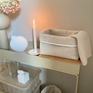 Puede incluir: Una cesta de almacenamiento tejida beige con un borde blanco y una toalla beige drapeada sobre el borde. La cesta est&aacute; sentada en un estante de madera con una vela encendida y un jarr&oacute;n blanco en el estante detr&aacute;s.