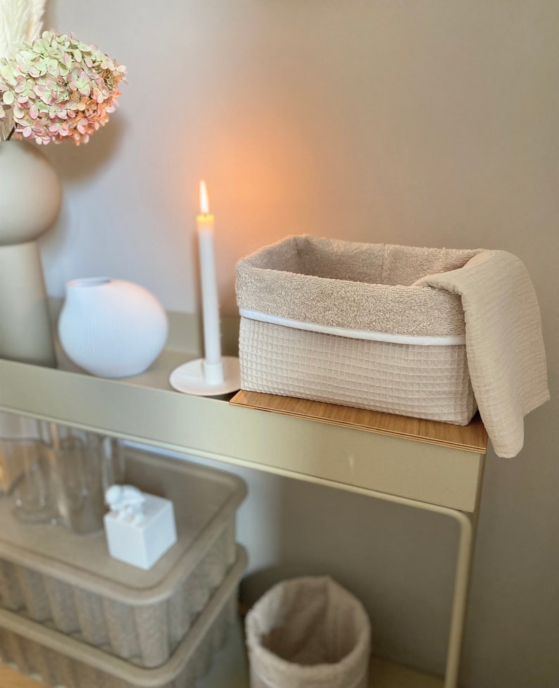 Puede incluir: Una cesta de almacenamiento tejida beige con un borde blanco y una toalla beige drapeada sobre el borde. La cesta est&aacute; sentada en un estante de madera con una vela encendida y un jarr&oacute;n blanco en el estante detr&aacute;s.