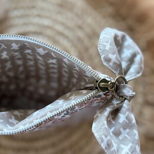 May include: A beige and white patterned cosmetic bag with a metal zipper and a decorative fabric bow. The interior lining features a repeating pattern of white shapes on a beige background. The bag's exterior has a floral pattern.