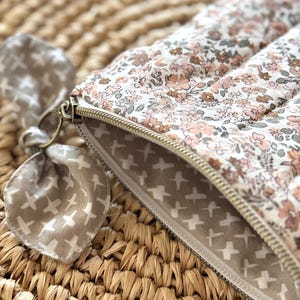 May include: A floral print quilted pouch with a gold zipper. The pouch features a pattern of small pink and brown flowers on a white background. A matching fabric bow is attached to the zipper pull. The interior lining has a pattern of white crosses on a tan background.