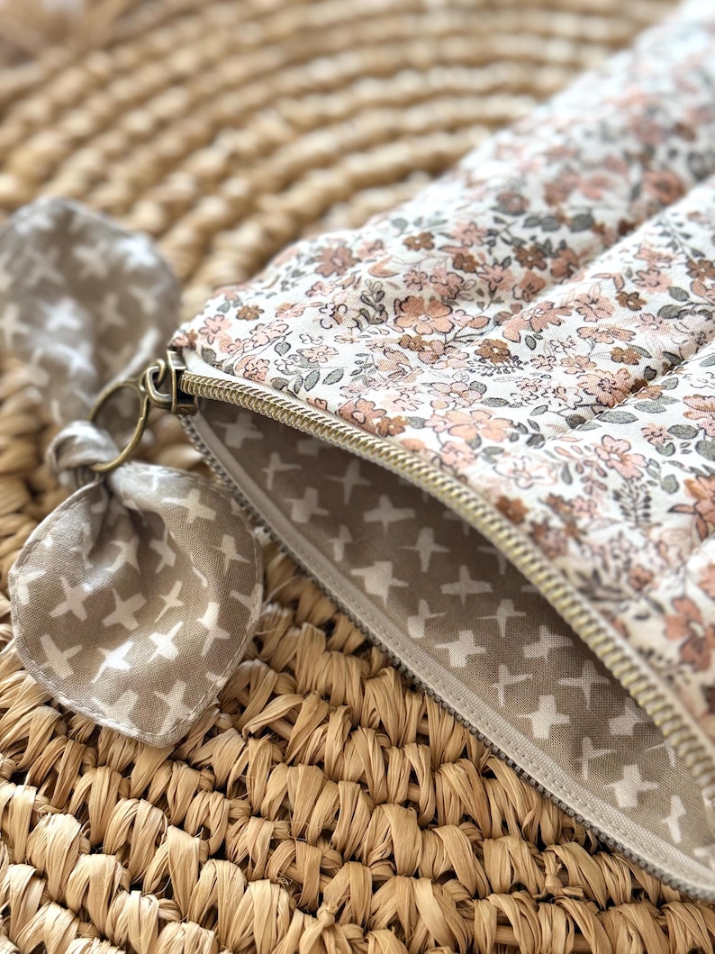 May include: A floral print quilted pouch with a gold zipper. The pouch features a pattern of small pink and brown flowers on a white background. A matching fabric bow is attached to the zipper pull. The interior lining has a pattern of white crosses on a tan background.