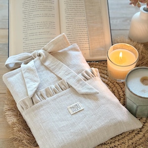 May include: A beige linen book sleeve with a ruffled pocket and bow closure, resting on a woven mat. An open book and a beige bookmark are in the background, along with a lit candle and a ceramic mug. The label on the sleeve reads "The Mini Pooco".
