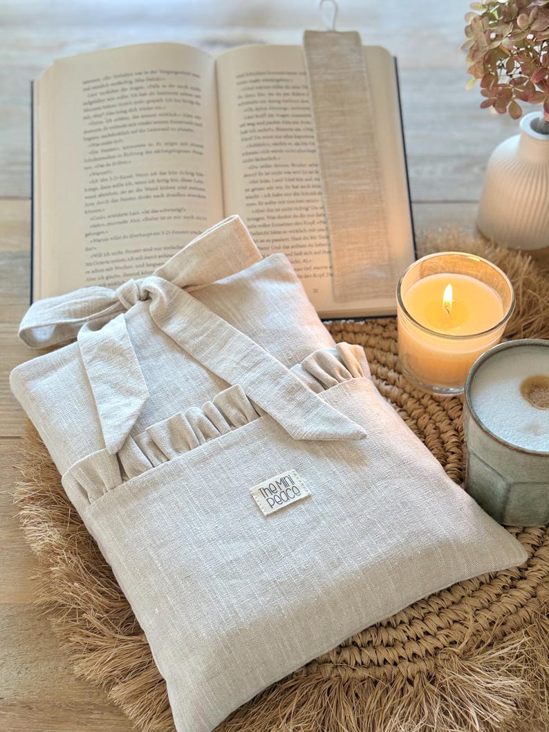 May include: A beige linen book sleeve with a ruffled pocket and bow closure, resting on a woven mat. An open book and a beige bookmark are in the background, along with a lit candle and a ceramic mug. The label on the sleeve reads "The Mini Pooco".