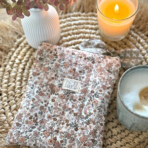 May include: A floral-patterned padded case with a tag that reads "The Mini Peace" sits on a woven mat. A lit candle in a glass, a white vase with dried flowers, and a mug with a foamy drink are also present.