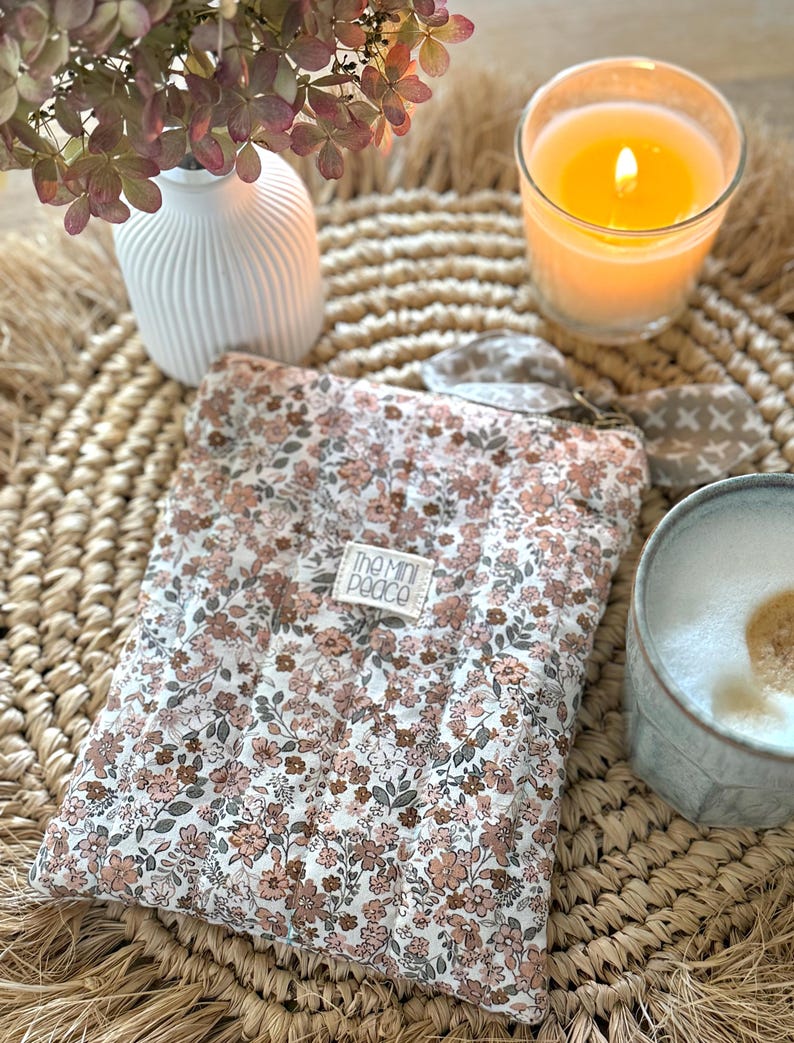 May include: A floral-patterned padded case with a tag that reads "The Mini Peace" sits on a woven mat. A lit candle in a glass, a white vase with dried flowers, and a mug with a foamy drink are also present.