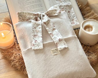 Peut inclure: Une housse de livre en velours côtelé beige avec un ruban floral et un marque-page assorti. La housse porte une petite étiquette avec l'inscription "The Mini Peace". Une bougie allumée et une tasse de café sont en arrière-plan.
