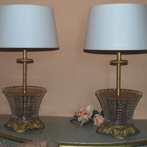May include: A pair of gold table lamps with white lampshades and crystal bases. The lamps are on a light blue table with a bouquet of pink flowers.