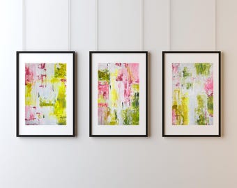 May include: Three framed abstract art prints. Each print features a colourful, textured design with pink, yellow, green, and white brushstrokes. The frames are black, and the art is displayed against a white wall.
