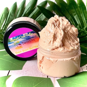 May include: A jar of shea body butter with a pink label that says "Sunset Over Belize Twinkles Shea Body Butter". The butter is a light brown color and has a whipped texture.