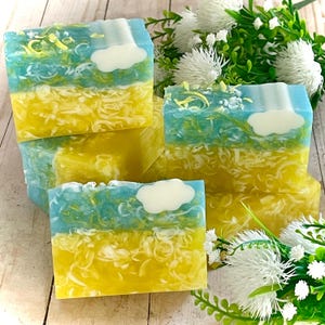 May include: Handmade soap bars with a layered design. The bars feature a yellow base, a blue and green middle, and a white cloud on top. The soap is decorated with small green and yellow accents. The soaps are arranged with white flowers.
