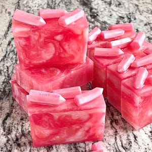 Pode incluir: V&aacute;rias barras de sabonete artesanal em tons de rosa e branco, com um padr&atilde;o marmorizado. Cada barra &eacute; coberta com pequenos enfeites em forma de cristal rosa claro. Os sabonetes est&atilde;o dispostos sobre uma superf&iacute;cie salpicada.