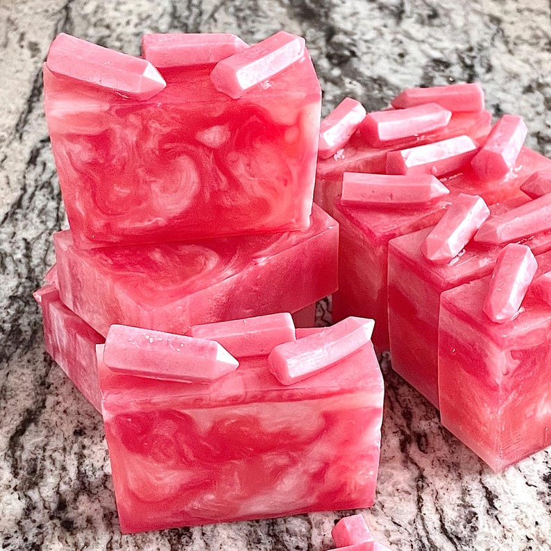 Pode incluir: V&aacute;rias barras de sabonete artesanal em tons de rosa e branco, com um padr&atilde;o marmorizado. Cada barra &eacute; coberta com pequenos enfeites em forma de cristal rosa claro. Os sabonetes est&atilde;o dispostos sobre uma superf&iacute;cie salpicada.