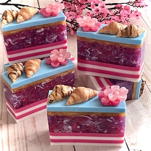 May include: A stack of decorative soaps designed to resemble miniature cakes. Each soap features a blue top, a layer of gold, a purple and white marbled center, and pink and white stripes. The soaps are topped with croissant and flower-shaped embellishments.