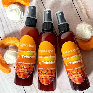 May include: Three 4 ounce bottles of peach and cream scented fragrance spray with a peach and cream colored label that says "Fragrance Spray Peaches and Cream Twinkles" and a website address. The bottles are sitting on a wooden surface with peach slices and white meringue cookies.