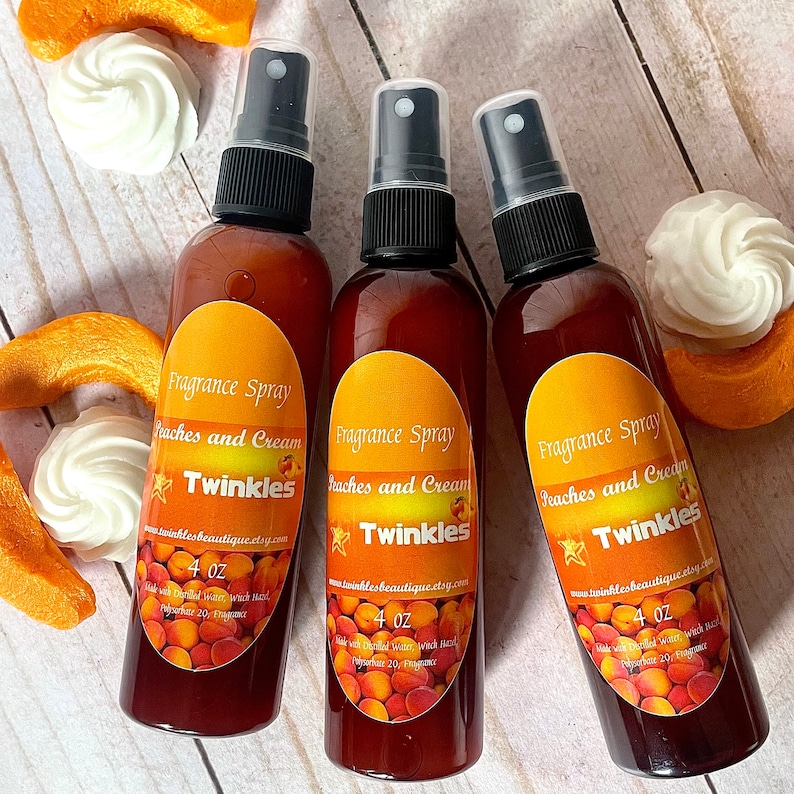 May include: Three 4 ounce bottles of peach and cream scented fragrance spray with a peach and cream colored label that says "Fragrance Spray Peaches and Cream Twinkles" and a website address. The bottles are sitting on a wooden surface with peach slices and white meringue cookies.