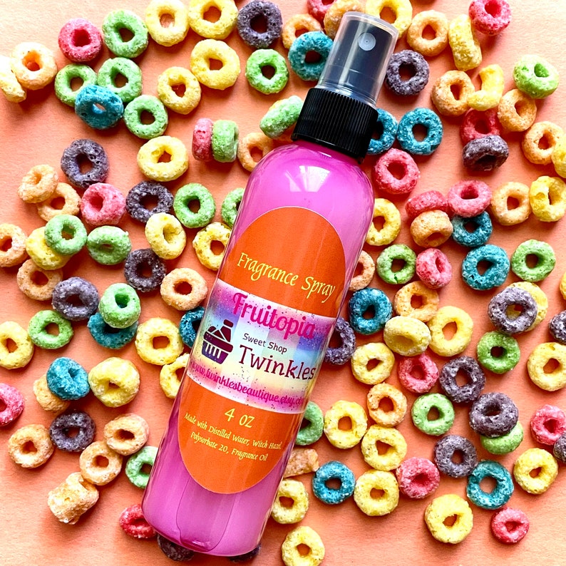 May include: A pink 4 oz bottle of Fruitotopia Sweet Shop Twinkles fragrance spray. The bottle is surrounded by colorful cereal rings on a peach-colored background. The label says "Fruitotopia Sweet Shop Twinkles" and "Made with Distilled Water, Witch Hazel, Polysorbate 20, Fragrance Oil".