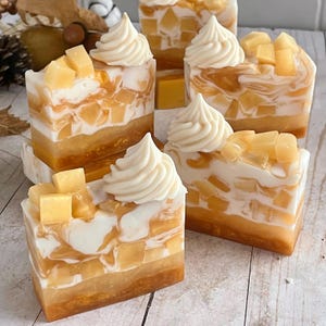 May include: Rectangular bars of soap featuring layers of translucent amber and opaque white swirls. Each bar is topped with a piped white soap dollop and embedded with small, square, translucent yellow soap cubes resembling fruit pieces.